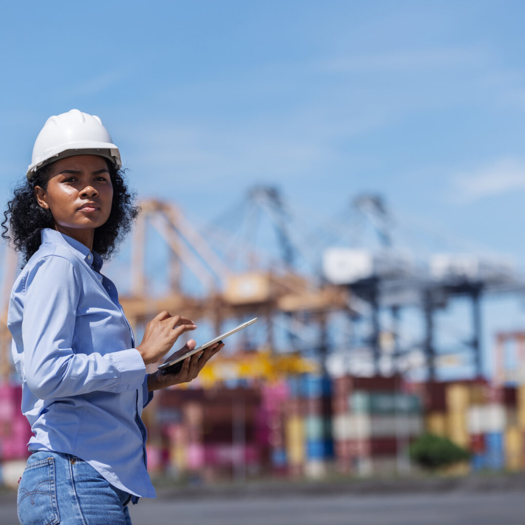 Woman in a hard hat using a tablet at a shipping port, representing business resilience and operational visibility amid global trade challenges.