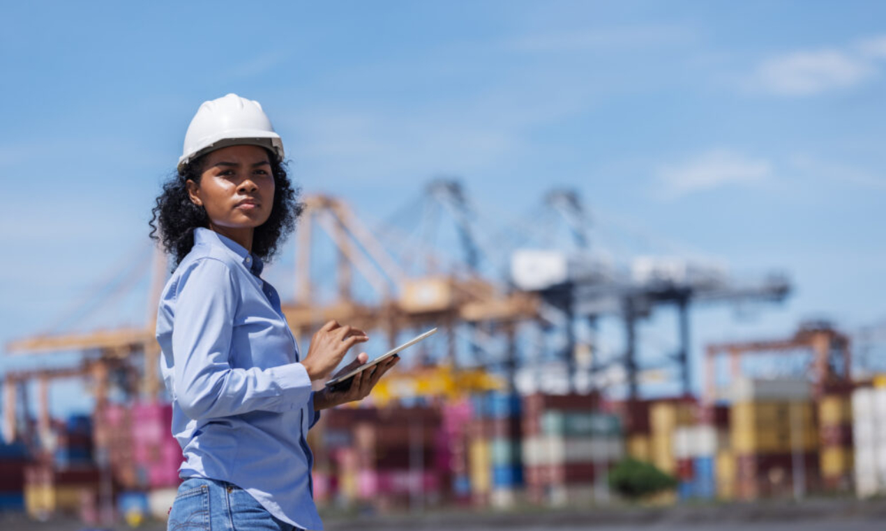 Woman in a hard hat using a tablet at a shipping port, representing business resilience and operational visibility amid global trade challenges.