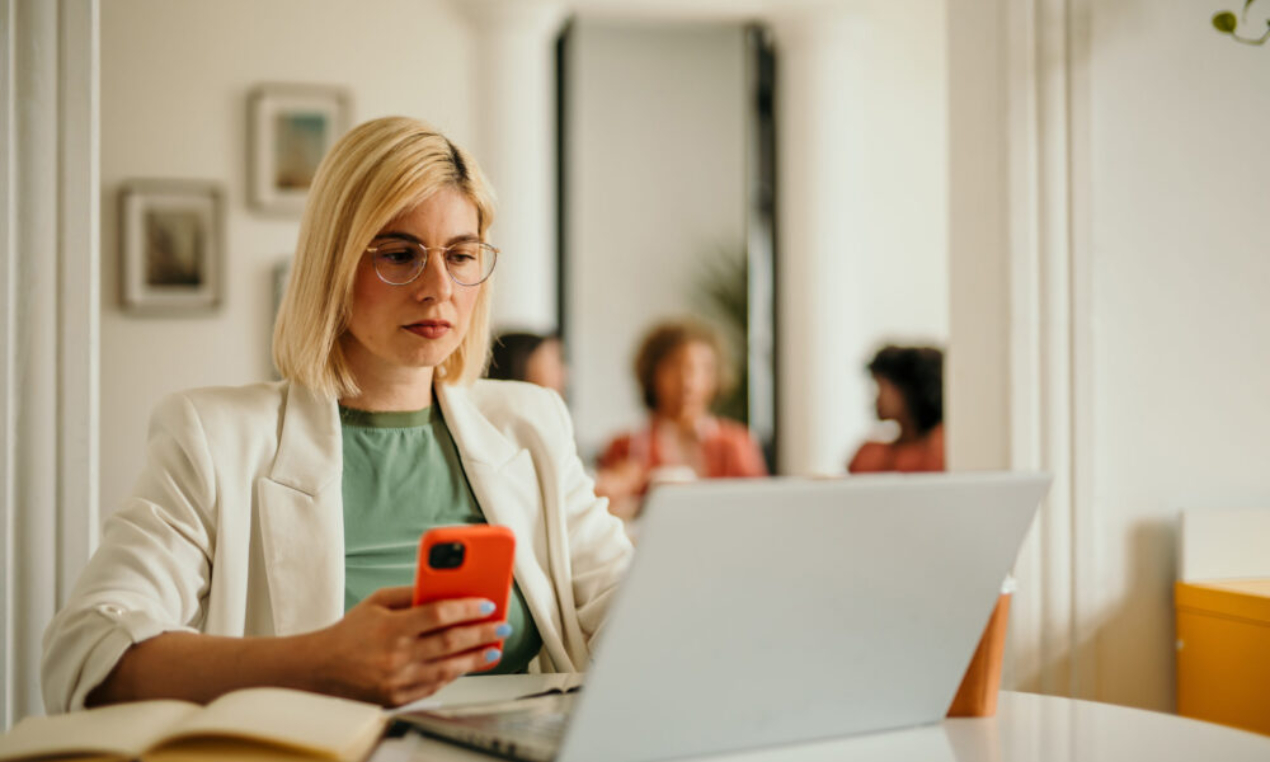 Businesswoman concentrating on her smartphone and laptop, working in a busy office environment with colleagues collaborating in the background