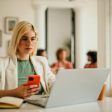 Businesswoman concentrating on her smartphone and laptop, working in a busy office environment with colleagues collaborating in the background