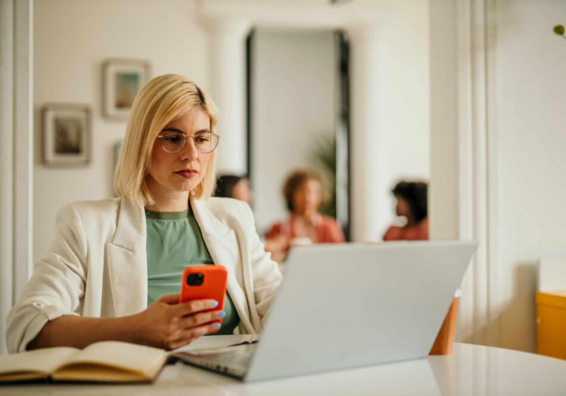 Businesswoman concentrating on her smartphone and laptop, working in a busy office environment with colleagues collaborating in the background